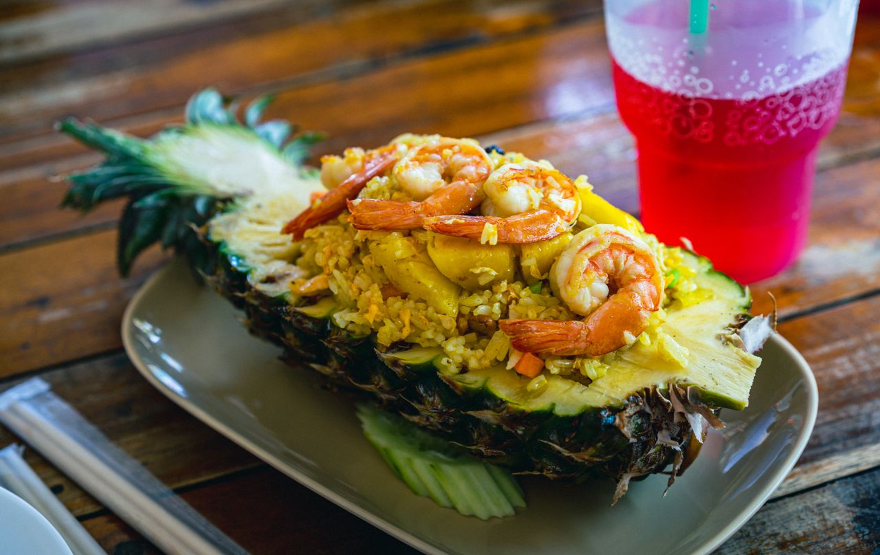 Delicious Thai pineapple fried rice with shrimp served in a pineapple shell with a red soda drink on the side.